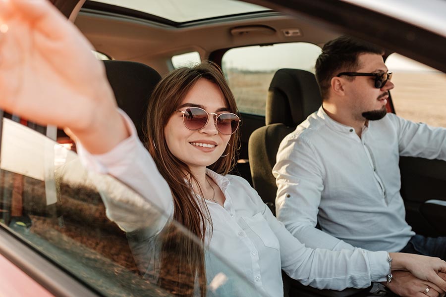 Blog - Young Couple Enjoying a Ride in a Red Car, Woman Waving Her Hand Out the Window